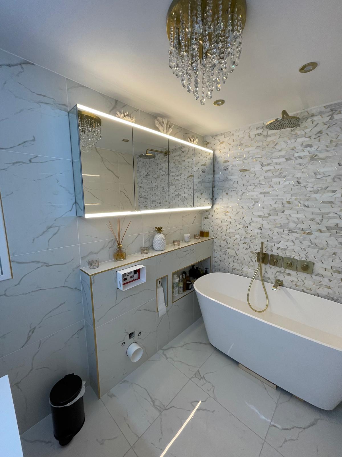 Marble bathroom — freestanding tub with gold rainfall against a mosaic feature wall, LED mirror and chandelier overhead